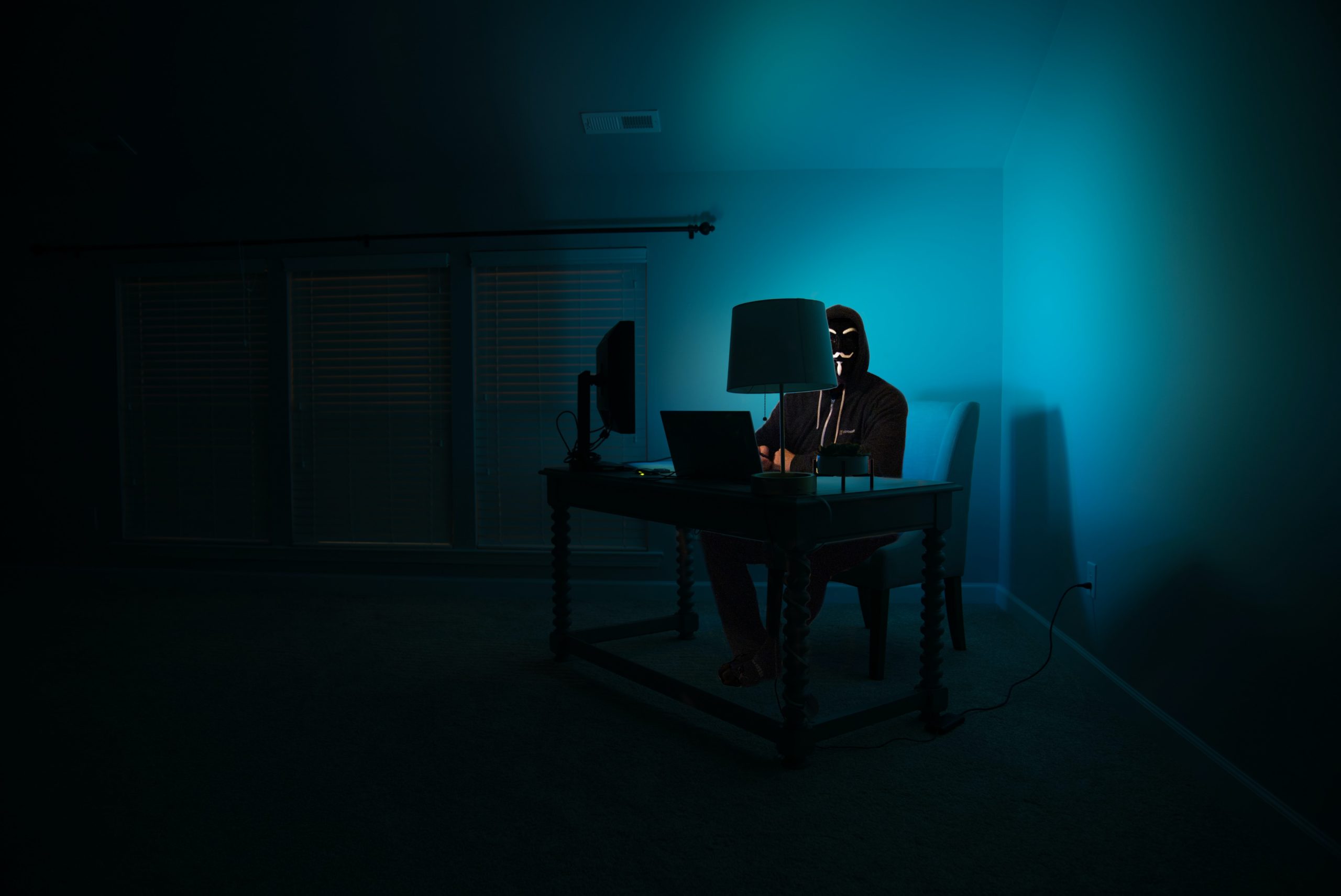 Person in a dark room on a computer wearing a face covering to hide their identity