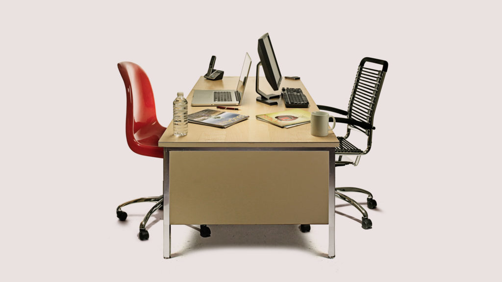 Desk with two chairs on either side with computers, books, a phone and coffee mug on the table.