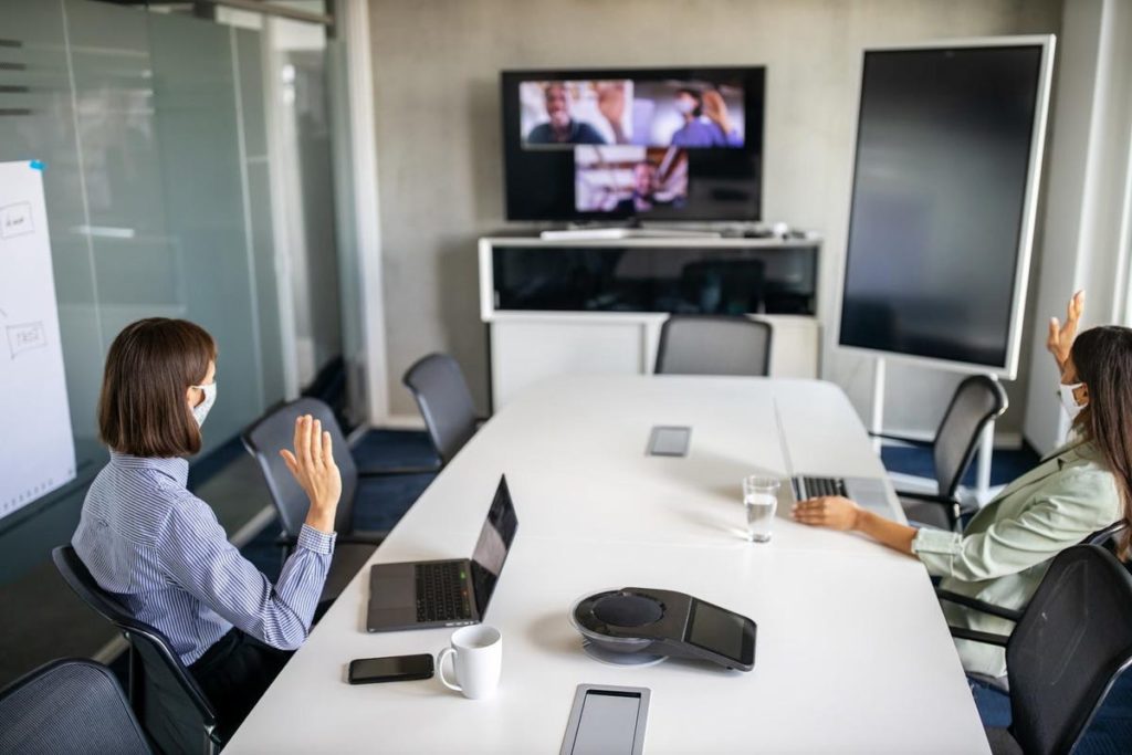 Two women in a meeting room waving to a video conference call